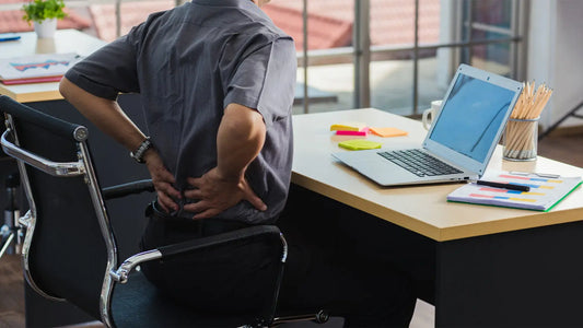 Office worker holding lower back in pain while sitting at a desk with a laptop, showing effects of poor posture and lack of lumbar support.